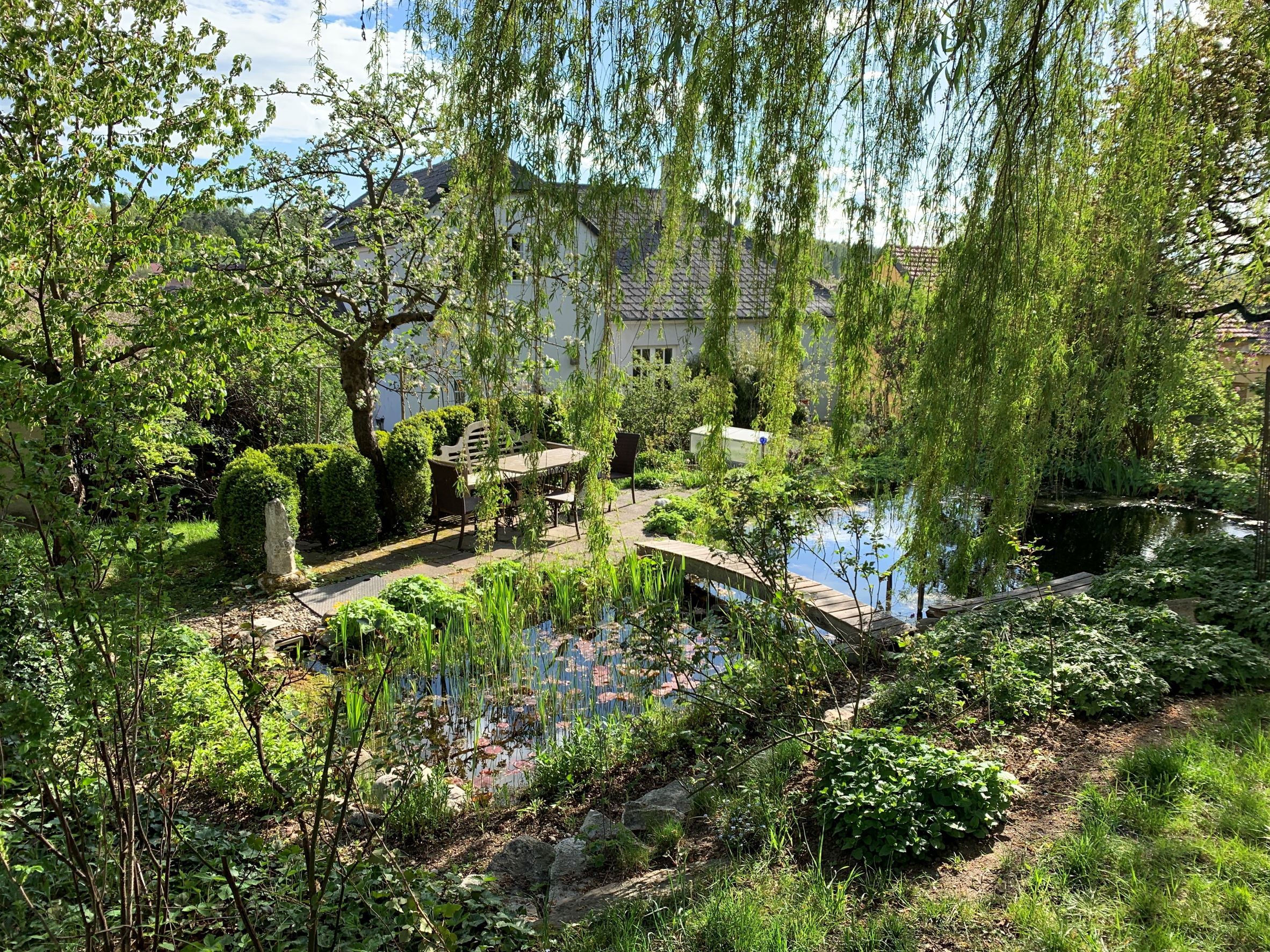 Ein idyllischer Garten mit Teich, Bäumen und einem Tisch mit Stühlen unter freiem Himmel.