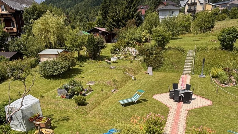 Ein gepflegter Garten mit Rasen, Gartenm&ouml;beln und einem Weg aus Pflastersteinen. Im Hintergrund sind H&auml;user und bewaldete H&uuml;gel zu sehen.