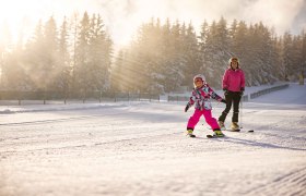 Kind und Erwachsener beim Skifahren im Schnee, umgeben von B&auml;umen und Sonnenstrahlen.