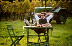Ein Mann entspannt sich an einem Tisch im Weinberg mit Wein, Brot und Äpfeln. Im Hintergrund steht ein grüner Traktor.