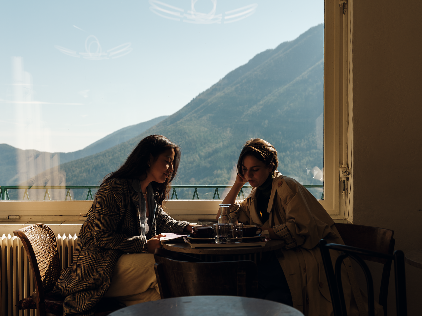 In der gemütlichen Atmosphäre der Kaffeehalle Semmering genießen zwei Freundinnen einen entspannten Moment mit Blick auf die majestätischen Berge. Das sanfte Licht, das durch die großen Fenster strömt, schafft eine einladende Stimmung, während sie sich über ihre Erlebnisse austauschen.
