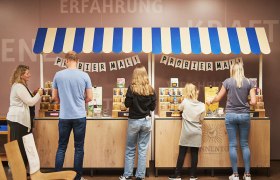 Menschen probieren Produkte an einem Stand mit blau-wei&szlig;em Dach.