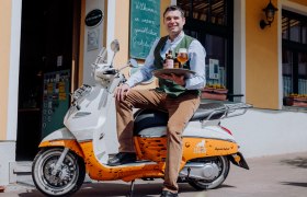 Ein Mann sitzt auf einem Roller vor einem Gebäude und hält ein Tablett mit Bierflaschen und einem Glas.