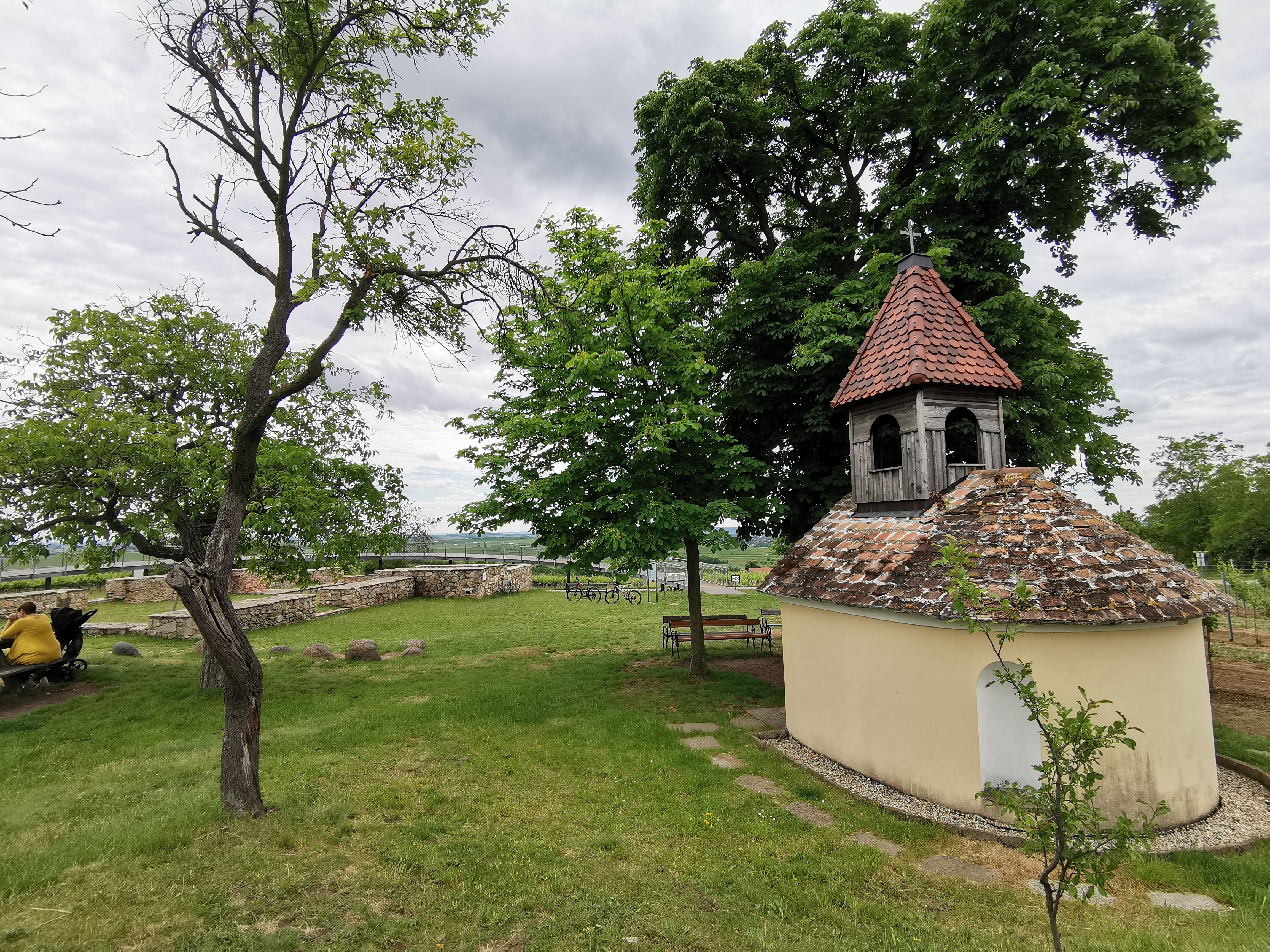 Kleine Kapelle mit rotem Ziegeldach in einer grünen Landschaft, umgeben von Bäumen und Bänken.