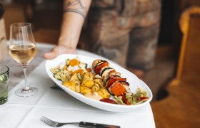 Ein Teller mit Grillspie&szlig;, Gem&uuml;se und Kartoffeln wird serviert, daneben ein Glas Wei&szlig;wein.