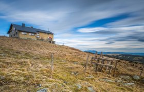 Das Wetterkoglerhaus auf einem Hügel mit bewölktem Himmel im Hintergrund.