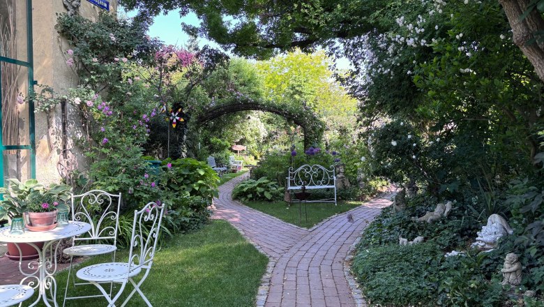 Ein idyllischer Garten mit wei&szlig;en Gartenm&ouml;beln, einem gepflasterten Weg und &uuml;ppiger Vegetation.