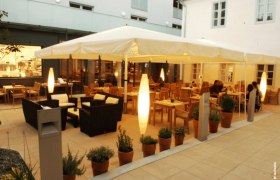 Ein beleuchteter Gastgarten mit Tischen, Stühlen und Pflanzen in Töpfen, umgeben von Gebäuden.