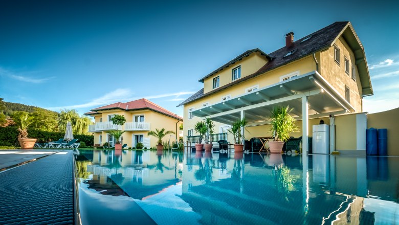 Die Gartenvilla mit gro&szlig;em Pool im Vordergrund, umgeben von Pflanzen und Liegest&uuml;hlen.