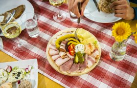 Ein gedeckter Tisch mit Aufschnittplatte, Brot, Wein und Blumen auf rot-wei&szlig; karierter Tischdecke.