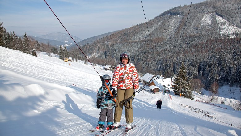 Zwei Skifahrer auf einem Schlepplift in einer verschneiten Berglandschaft.