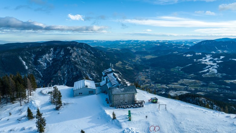 Luftaufnahme des Raxalm-Berggasthofs im Winter mit schneebedeckten Bergen und Tälern im Hintergrund.