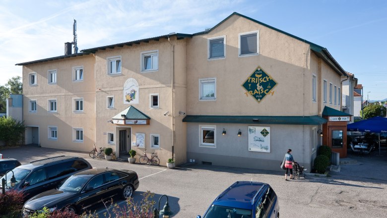 Außenansicht eines Gebäudes mit der Aufschrift 'Pension Schlafgut' und 'Café Trisch & Tratsch'. Autos parken davor, eine Person mit Kinderwagen geht vorbei.