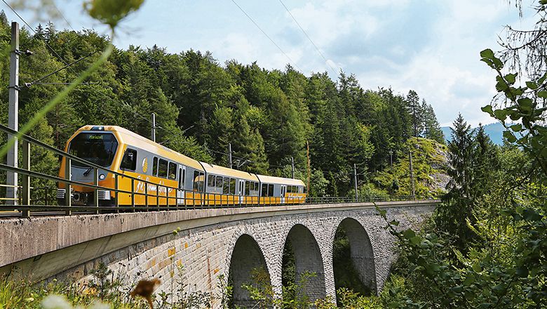 Ein gelber Zug der Mariazellerbahn fährt über eine Steinbrücke in einer bewaldeten Landschaft.
