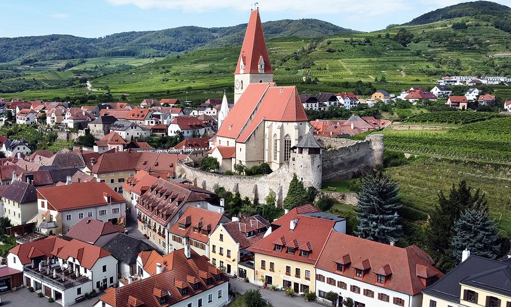 Luftaufnahme eines Dorfes mit Kirche und Weinbergen im Hintergrund.