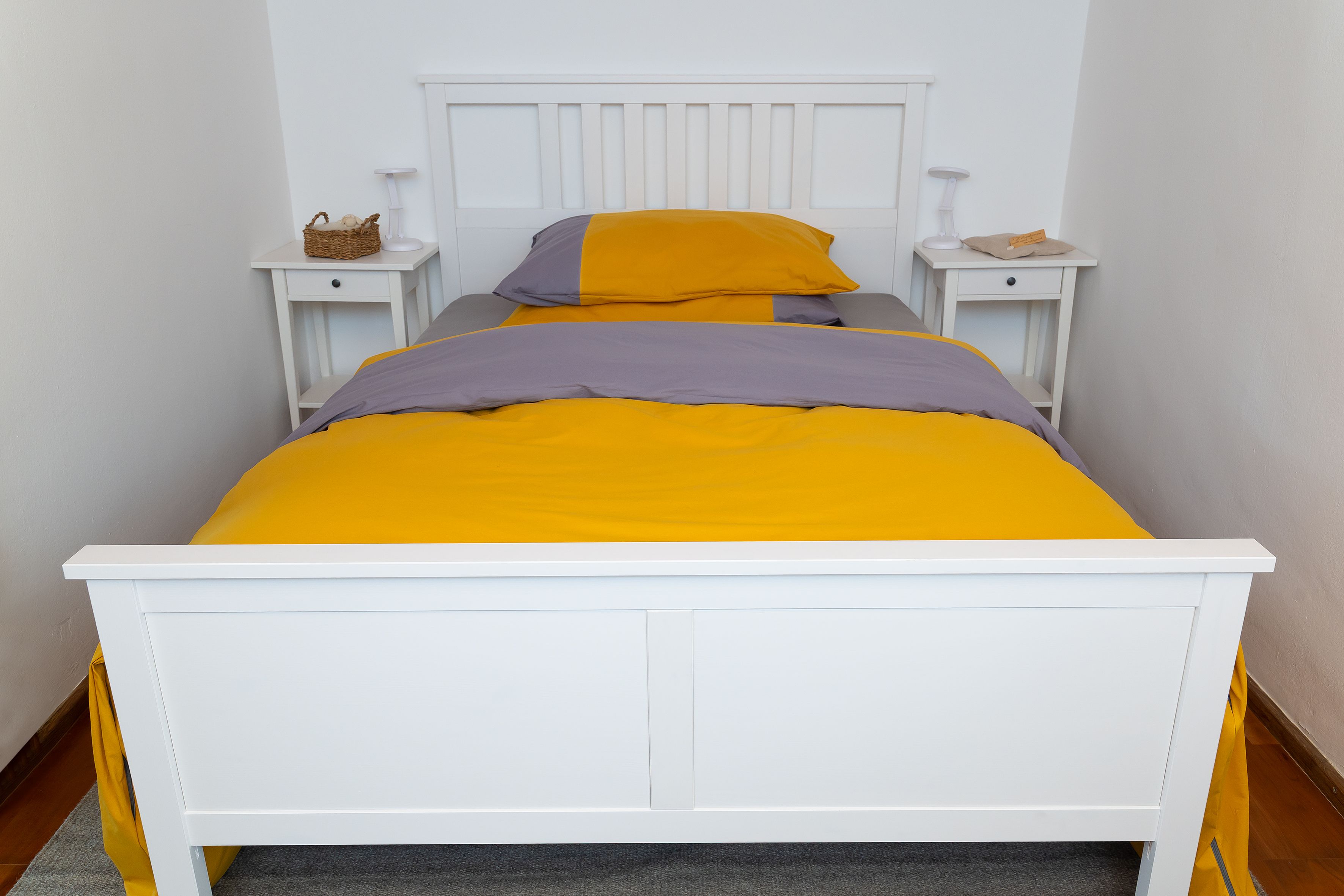 Ein weißes Bett mit gelb-grauer Bettwäsche in einem schlichten Schlafzimmer.