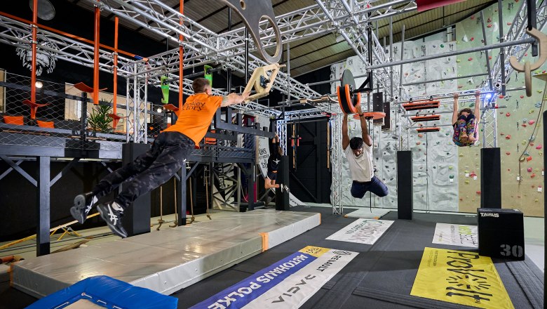 Menschen trainieren in einem Indoor-Kletterpark mit verschiedenen Hindernissen und Kletterw&auml;nden.
