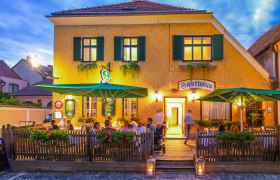 Außenansicht eines Gasthauses mit Terrasse und Gästen bei Abenddämmerung.