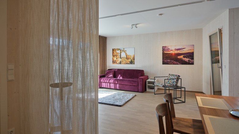 Wohnzimmer mit lila Sofa, Holztisch und St&uuml;hlen, Vorhang, und Wandbildern.