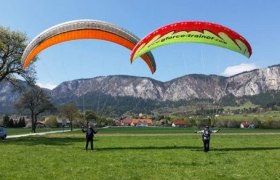 Zwei Personen beim Paragliding-Training auf einer Wiese vor einer Berglandschaft.