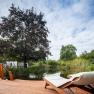 Holzterrasse mit Liegestühlen am Schwimmbiotop, umgeben von Bäumen und Pflanzen.