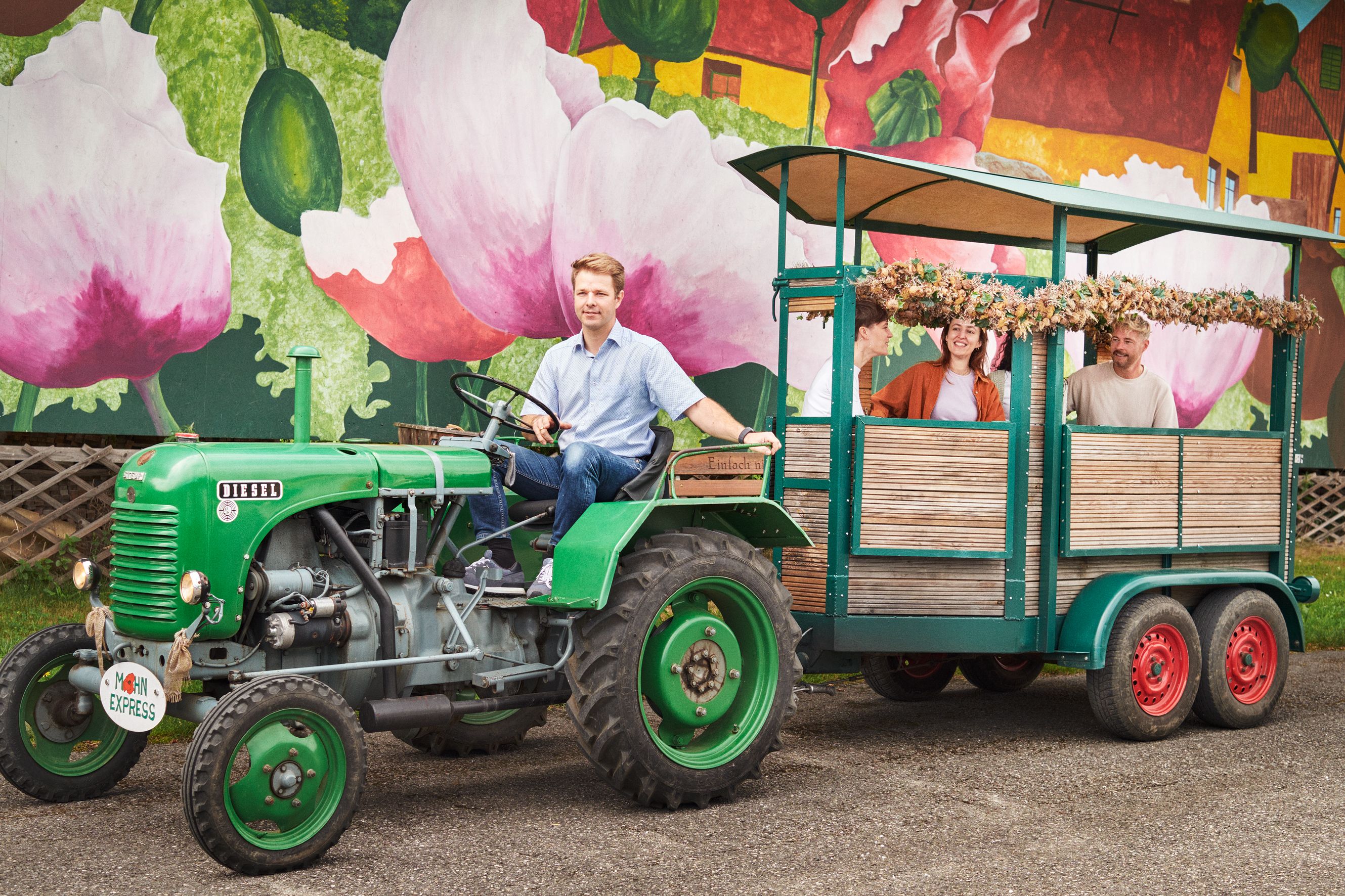 Ein grüner Traktor zieht einen Anhänger mit fröhlichen Menschen vor einer bunten Wand mit Mohnblumenmalerei.