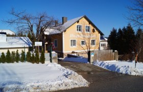 Ein gelbes Haus mit der Aufschrift 'Haus Bergblick' im Schnee, umgeben von B&auml;umen und einem blauen Himmel.