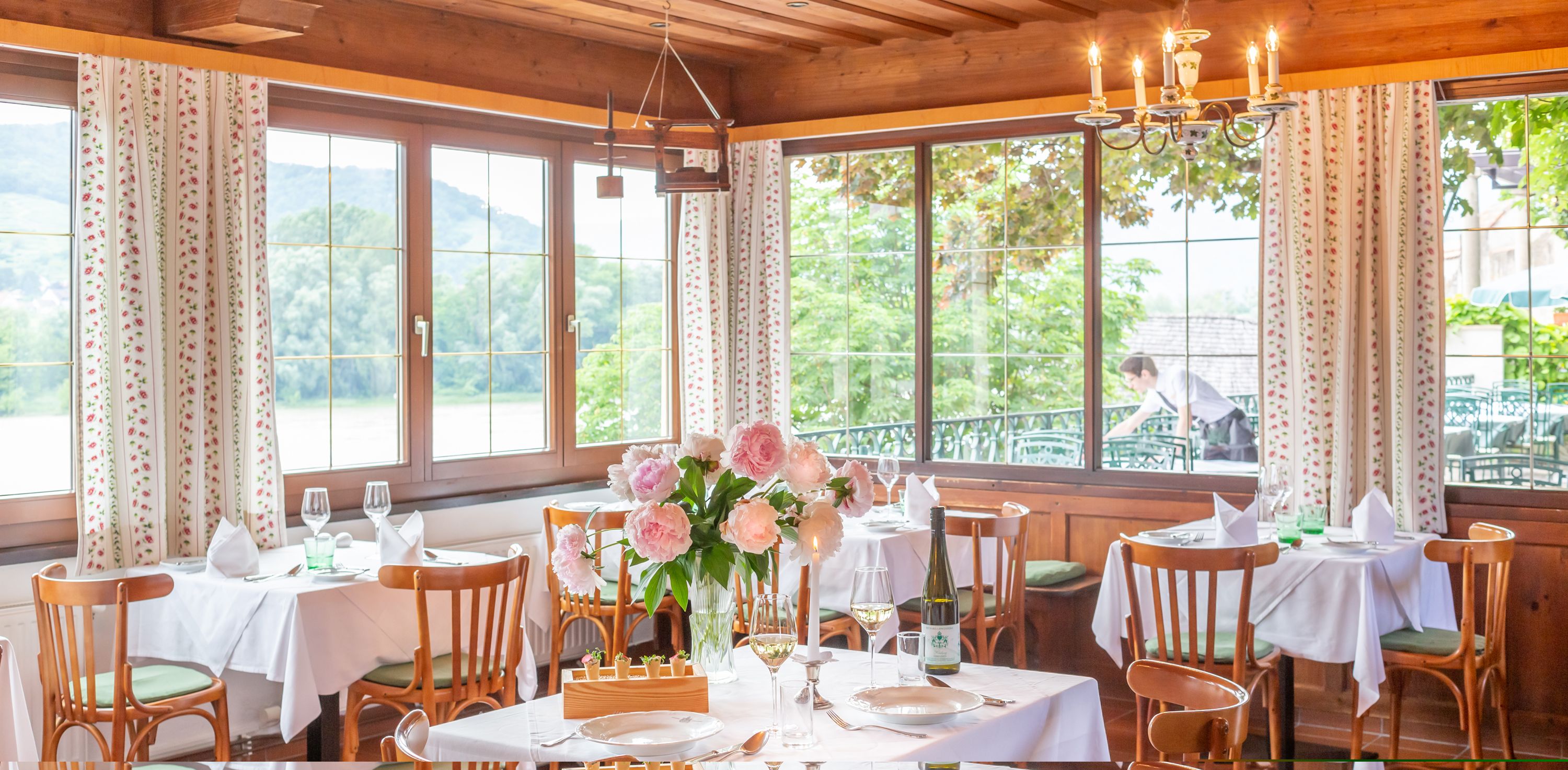 Elegantes Restaurant mit gedeckten Tischen, Blumen und Blick auf die Natur durch große Fenster.