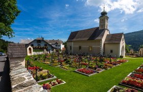 Pfarrkirche St. Peter am Neuwald mit gepflegtem Friedhof und blühenden Blumenbeeten, umgeben von grüner Landschaft und blauem Himmel.