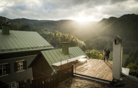 Zwei Personen stehen auf einer Terrasse mit Blick auf bewaldete Berge bei Sonnenuntergang.