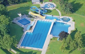 Luftaufnahme eines Freibads mit mehreren Becken und Wasserrutschen, umgeben von grünen Bäumen und Rasenflächen.