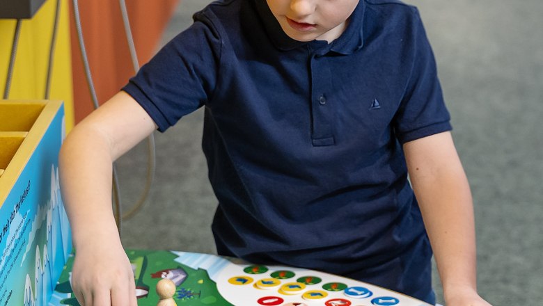 Ein Kind spielt ein Brettspiel mit bunten Spielfiguren und W&uuml;rfeln auf einem Tisch.