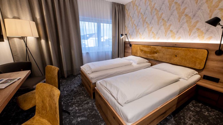 Modernes Gästezimmer mit zwei Einzelbetten, Holzmöbeln, gelben Stühlen und einer Stehlampe. Helle Vorhänge und gemusterte Tapete an der Wand.