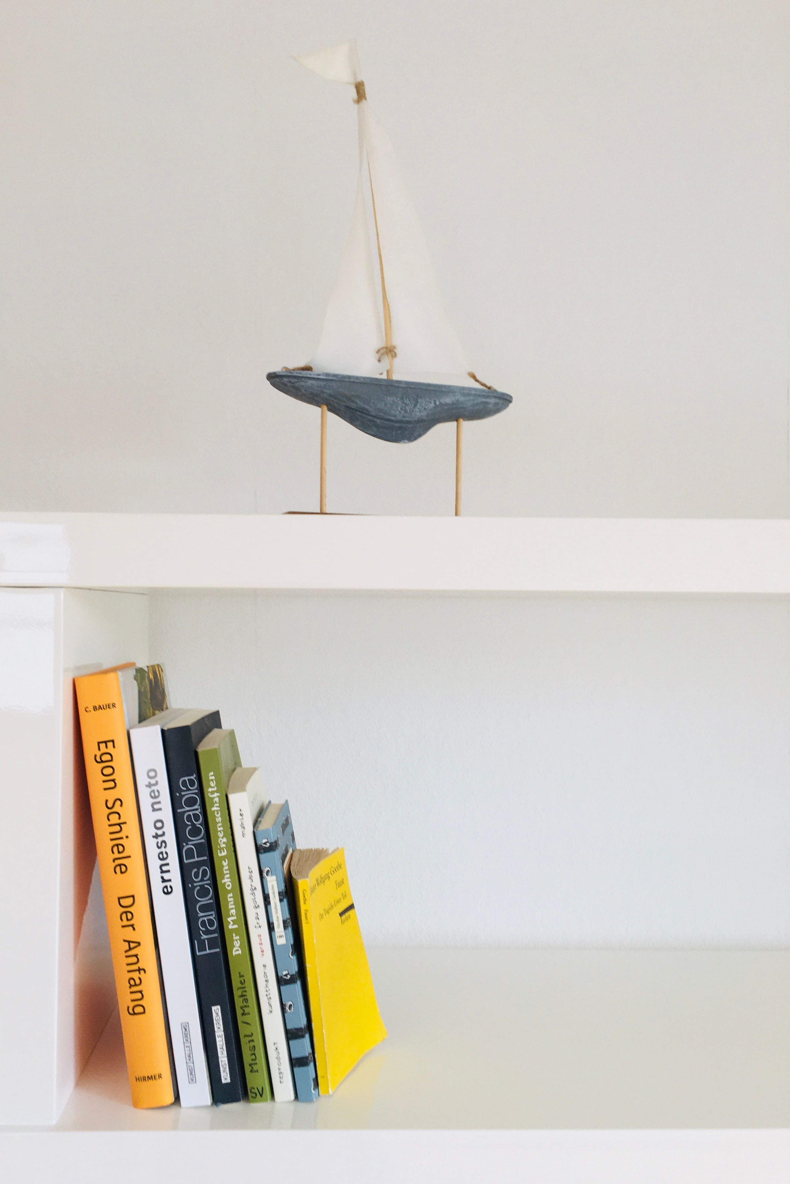 Ein weißes Regal mit Büchern und einem kleinen Segelboot als Dekoration.