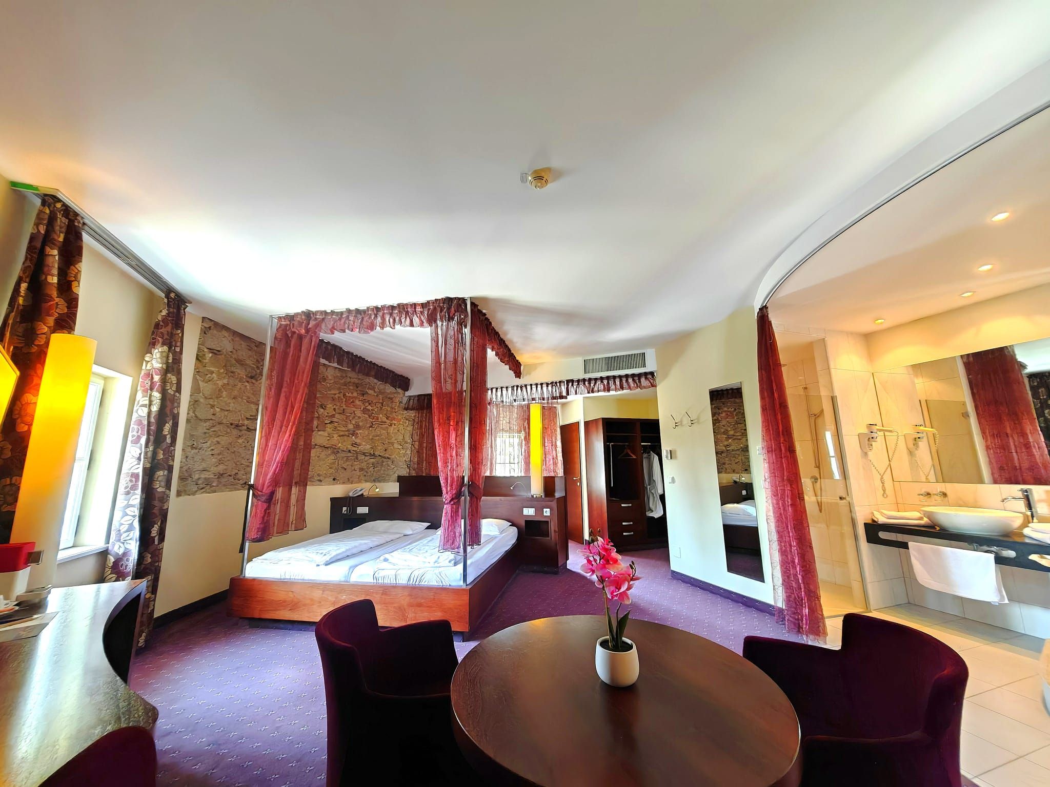 Ein elegantes Hotelzimmer mit einem Himmelbett, einem runden Tisch mit Blumen und einem offenen Badezimmerbereich.