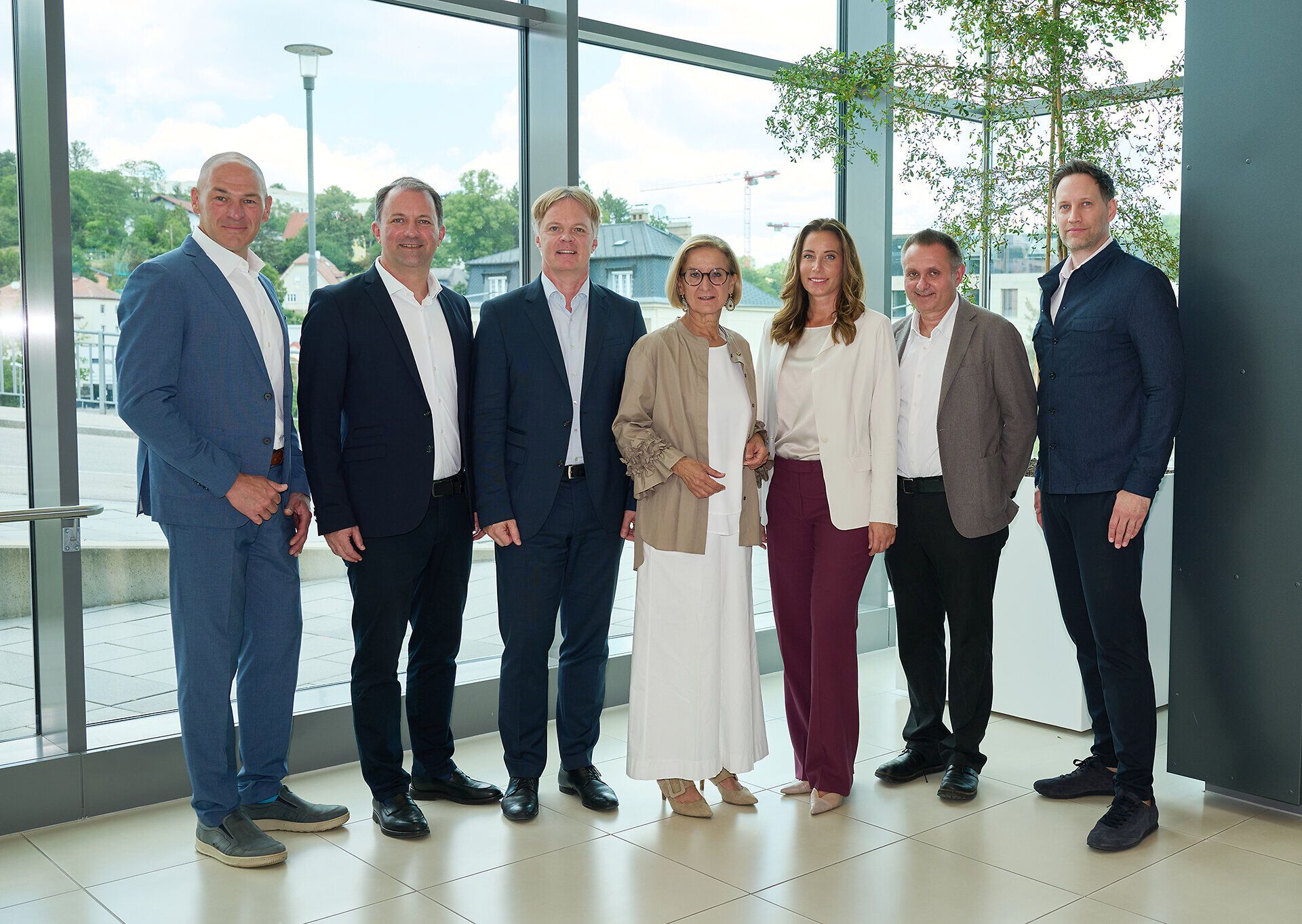 Sieben Personen stehen nebeneinander für ein Gruppenfoto in einem hellen, modernen Innenraum mit großen Fensterflächen. Im Hintergrund sind Gebäude, Bäume und eine Terrasse zu sehen.