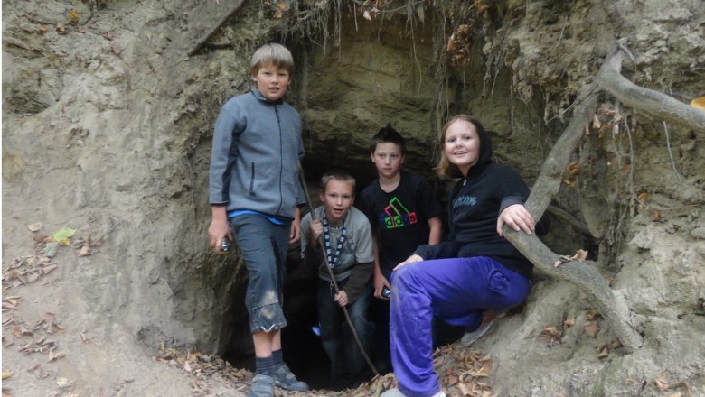 Vier Kinder stehen vor einem H&ouml;hleneingang in einem Wald.