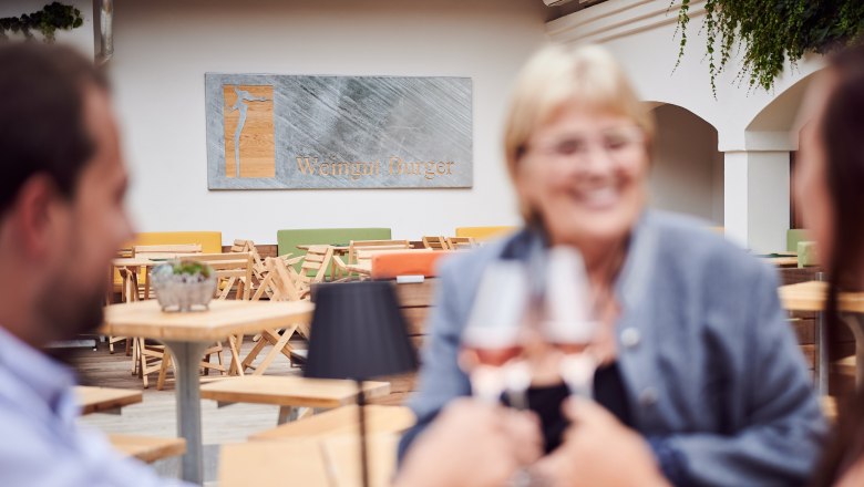 Drei Personen stoßen mit Weingläsern in einem Innenhof an, im Hintergrund ein Schild mit der Aufschrift 'Weingut Burger'.