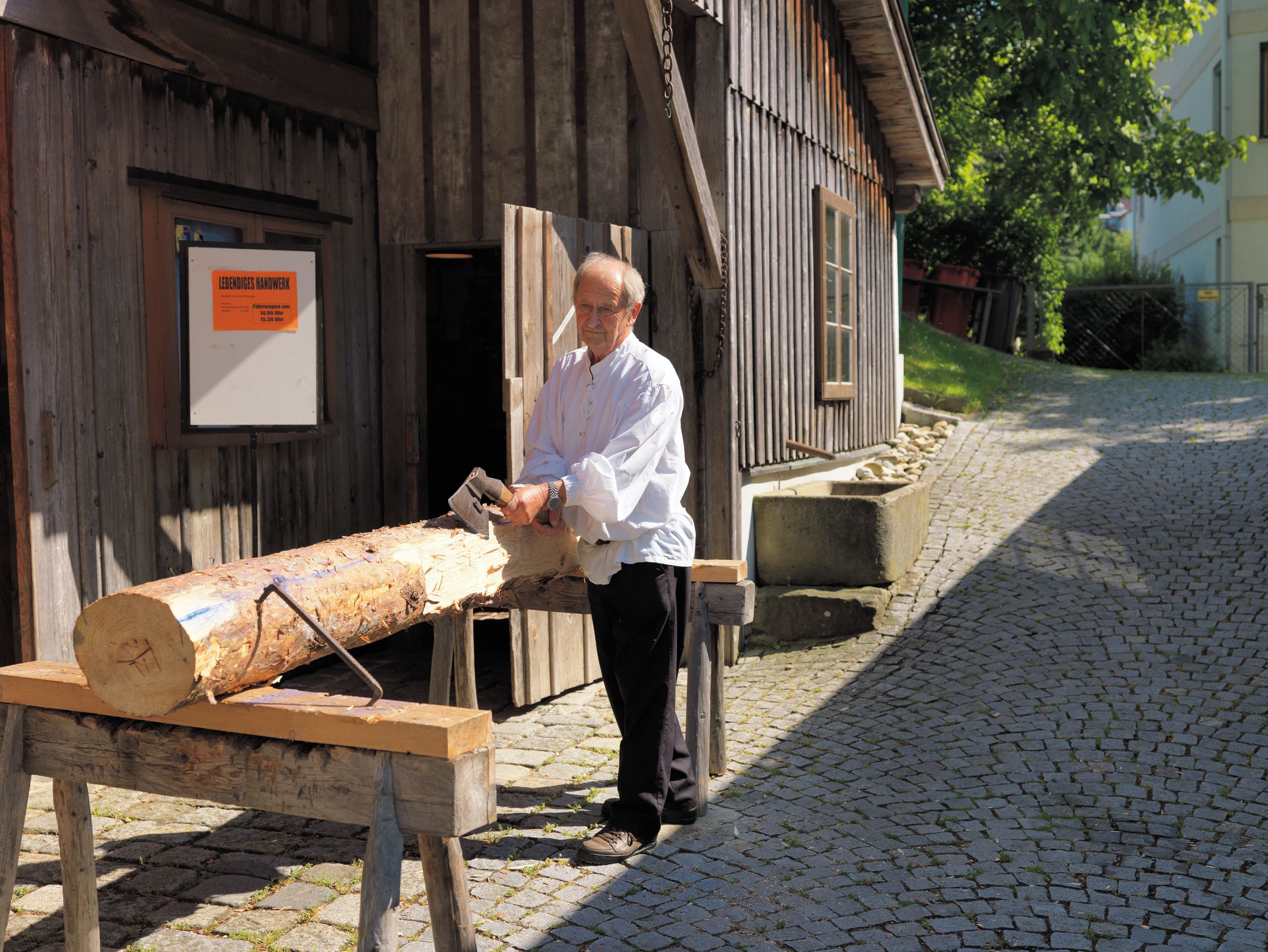 Ein Mann bearbeitet einen Baumstamm mit einer Axt vor einem Holzhaus.