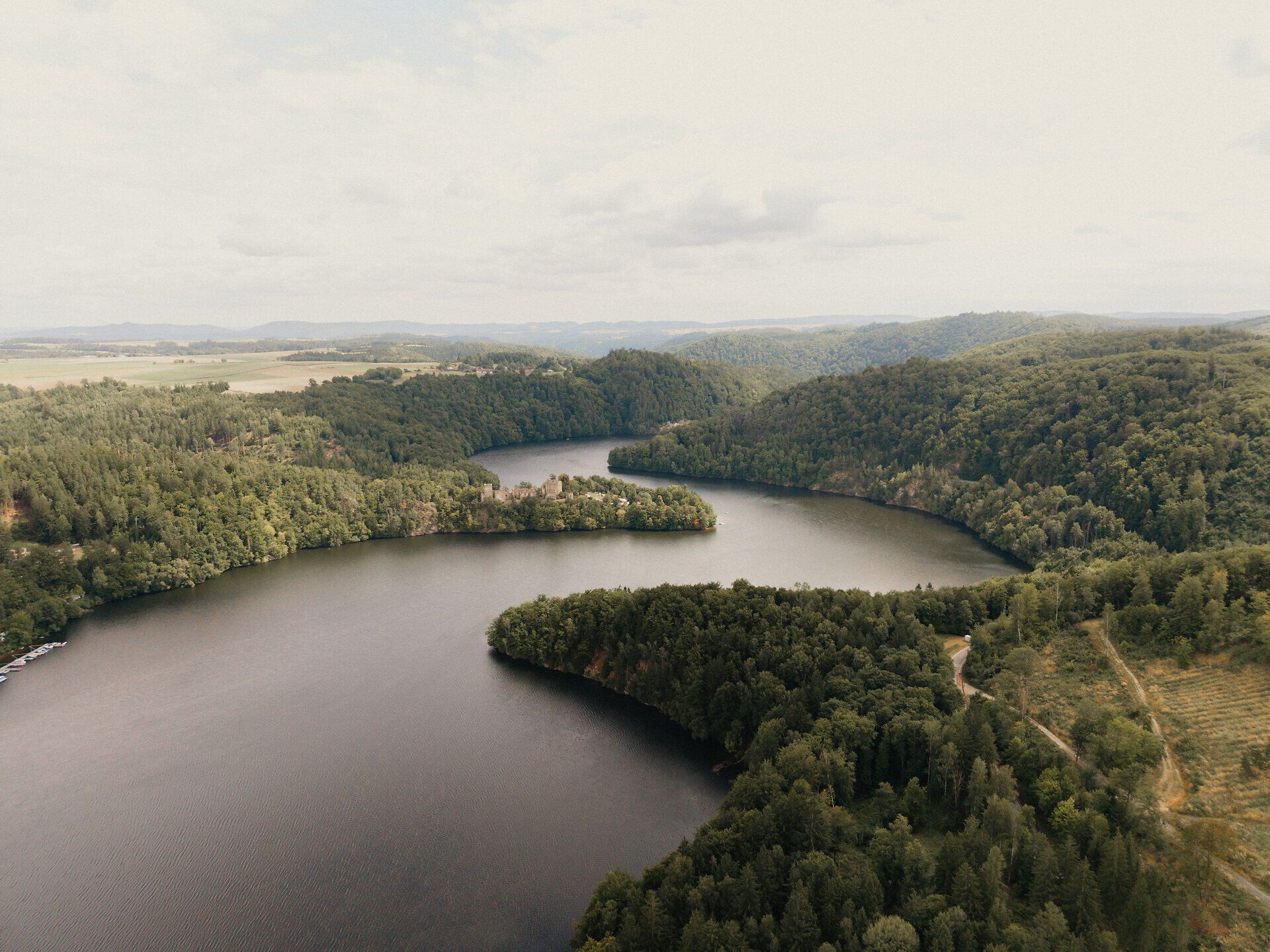 Luftaufnahme des Stausees Dobra im Waldviertel, mit sanft geschwungenem See, dicht bewaldeten Hügeln und weiter Landschaft unter hellem Himmel.