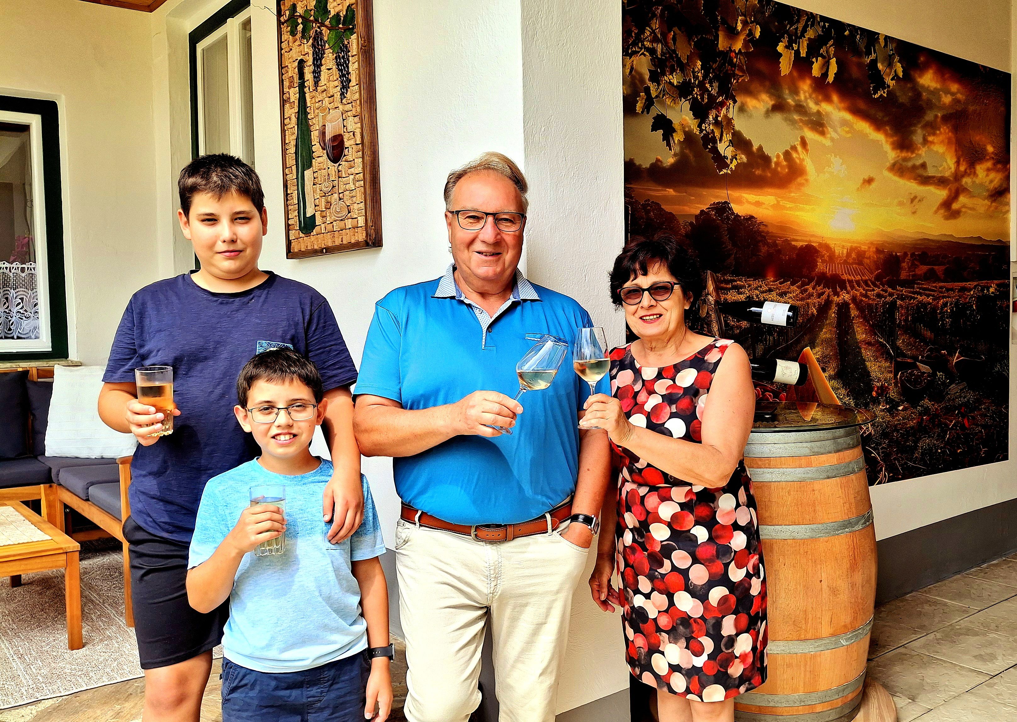 Eine Familie steht vor einem Weinfass und einem Bild von Weinbergen, hält Gläser und lächelt in die Kamera.