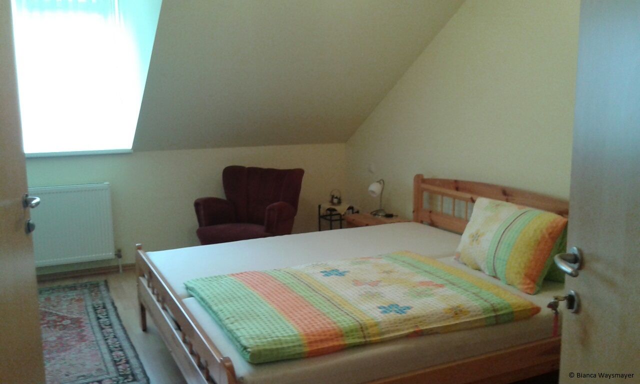 Ein gemütliches Schlafzimmer mit einem Doppelbett, einem roten Sessel und einem kleinen Nachttisch unter einer Dachschräge.