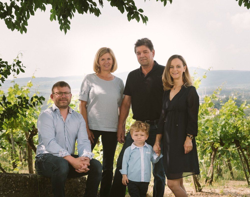 Fünf Personen posieren in einem Weinberg, darunter ein Kind. Im Hintergrund sind Hügel und Weinreben zu sehen.