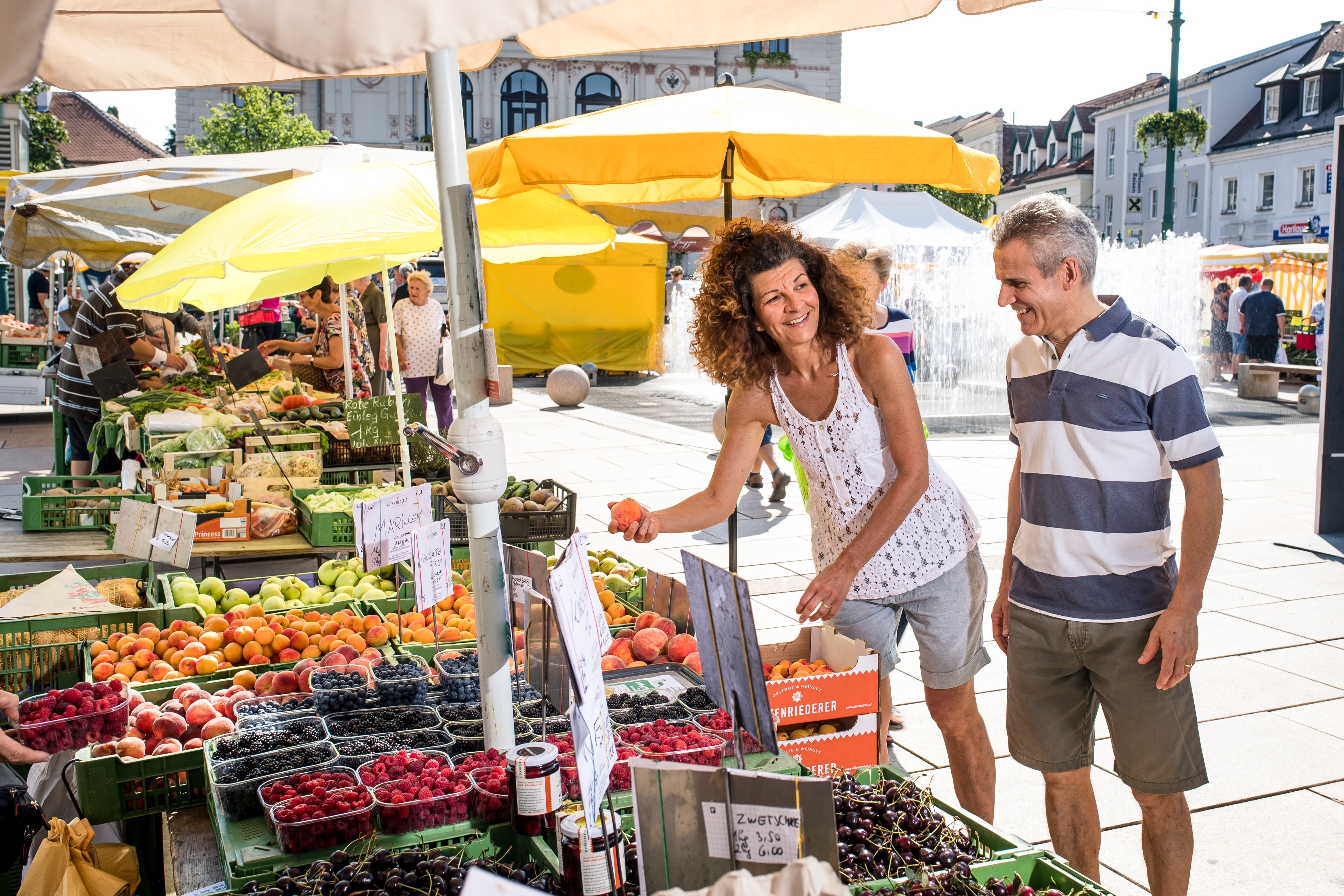 Menschen kaufen Obst auf einem Markt mit gelben Sonnenschirmen.
