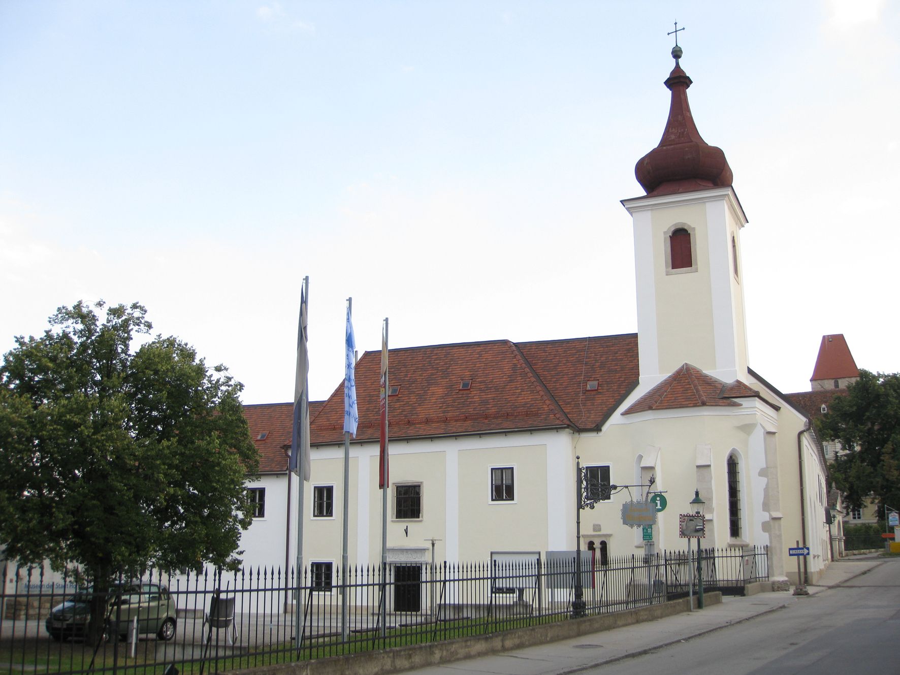Ein Gebäude mit Kirchturm und roten Dachziegeln, umgeben von einem Zaun und Bäumen.