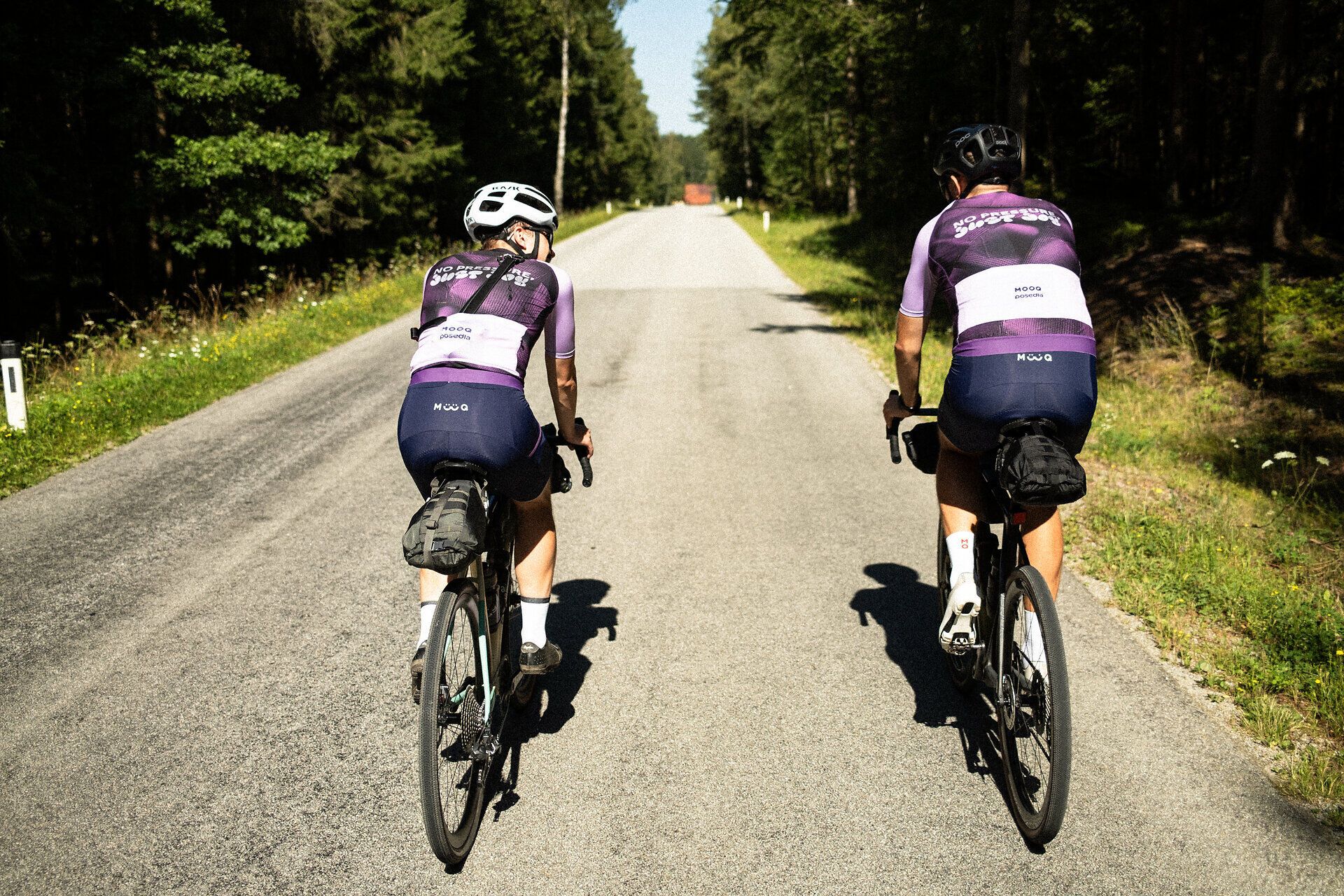 Zwei Radfahrer genießen die frische Luft und die ruhige Atmosphäre auf einer malerischen Landstraße, umgeben von üppigem Grün und hohen Bäumen. Die sanften Hügel im Hintergrund laden dazu ein, die Schönheit der Natur zu erkunden und die Freiheit des Radfahrens zu erleben.