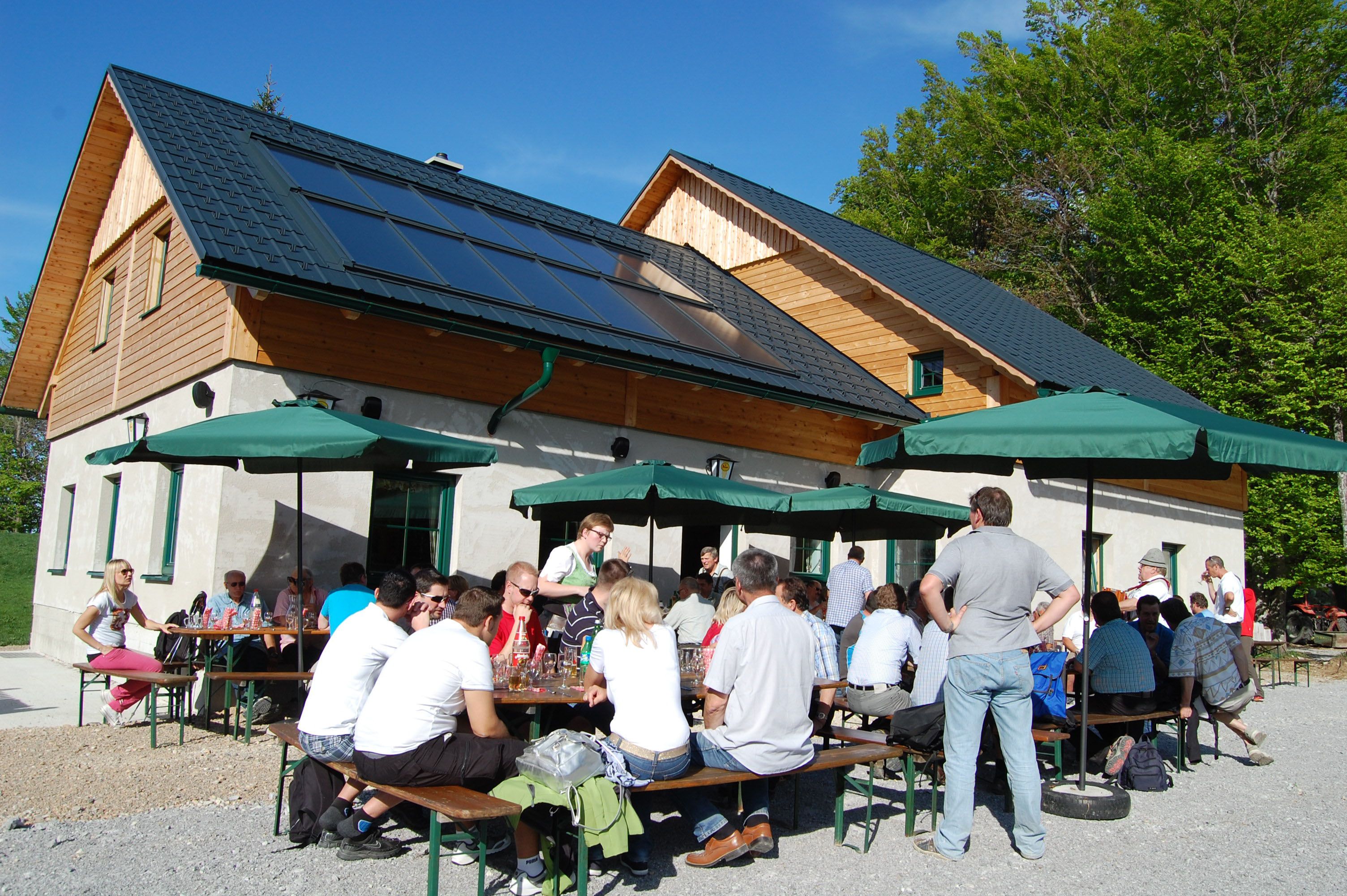 Menschen sitzen an Tischen vor einer Hütte mit Solarpanelen und Sonnenschirmen.