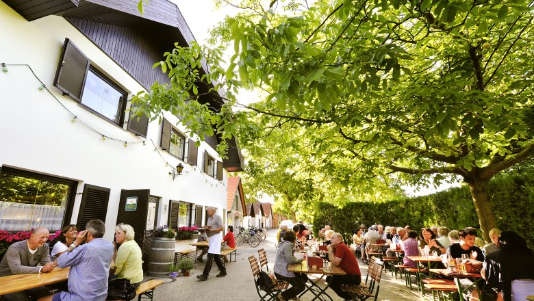 Menschen sitzen an Tischen im Freien unter B&auml;umen bei einem Heurigenbetrieb.