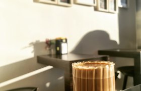 Ein Glas Kaffee auf einem Tisch in einem Café mit Bilderrahmen an der Wand im Hintergrund.