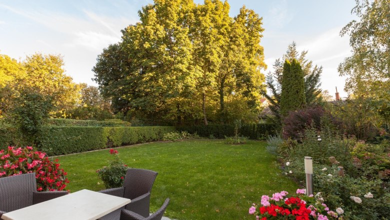 Ein gepflegter Garten mit Rasen, Blumenbeeten und Gartenm&ouml;beln auf einer Terrasse.